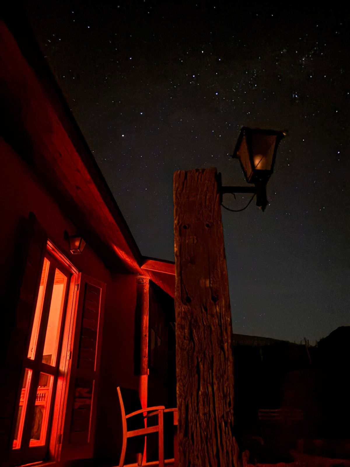 Céu estrelado sobre a varanda de um chalé, com o lampião e a luz quente da janela