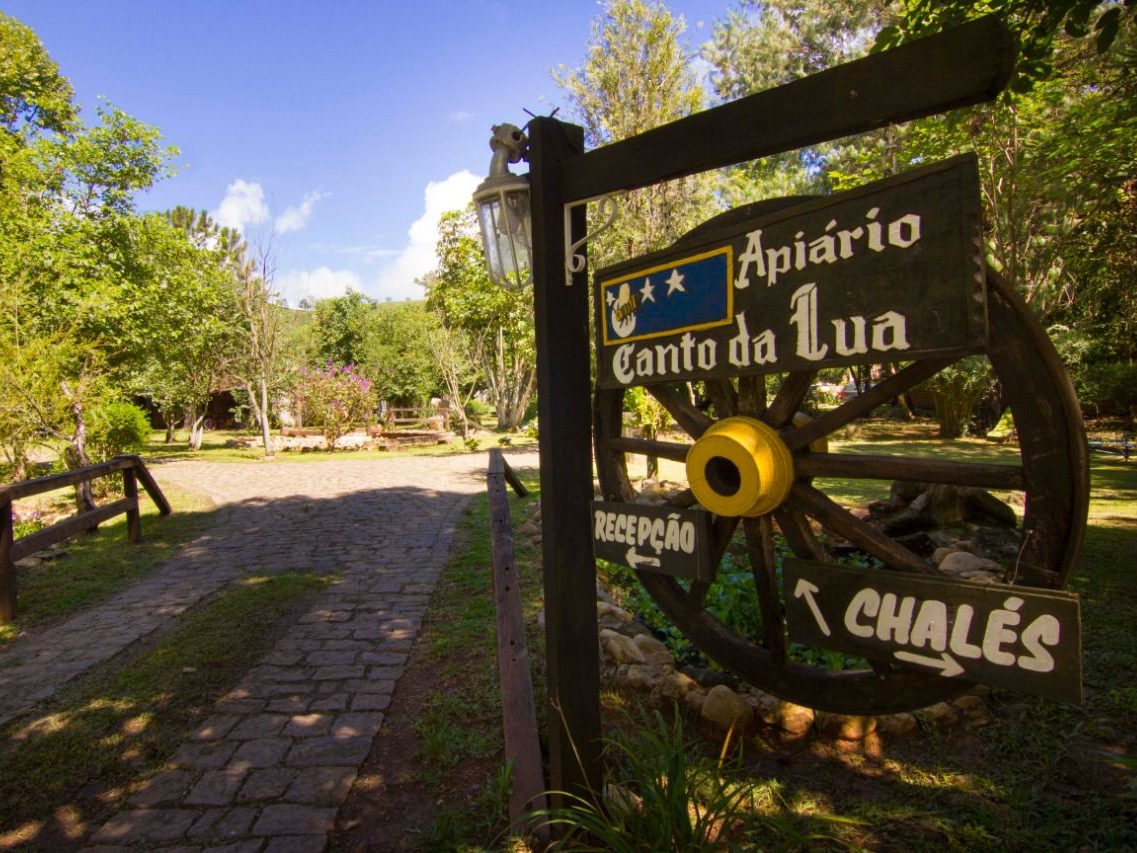 Placa de madeira do Canto da Lua na entrada, com setas para a Recepção e os Chalés, ao lado do caminho de pedra em dia de sol