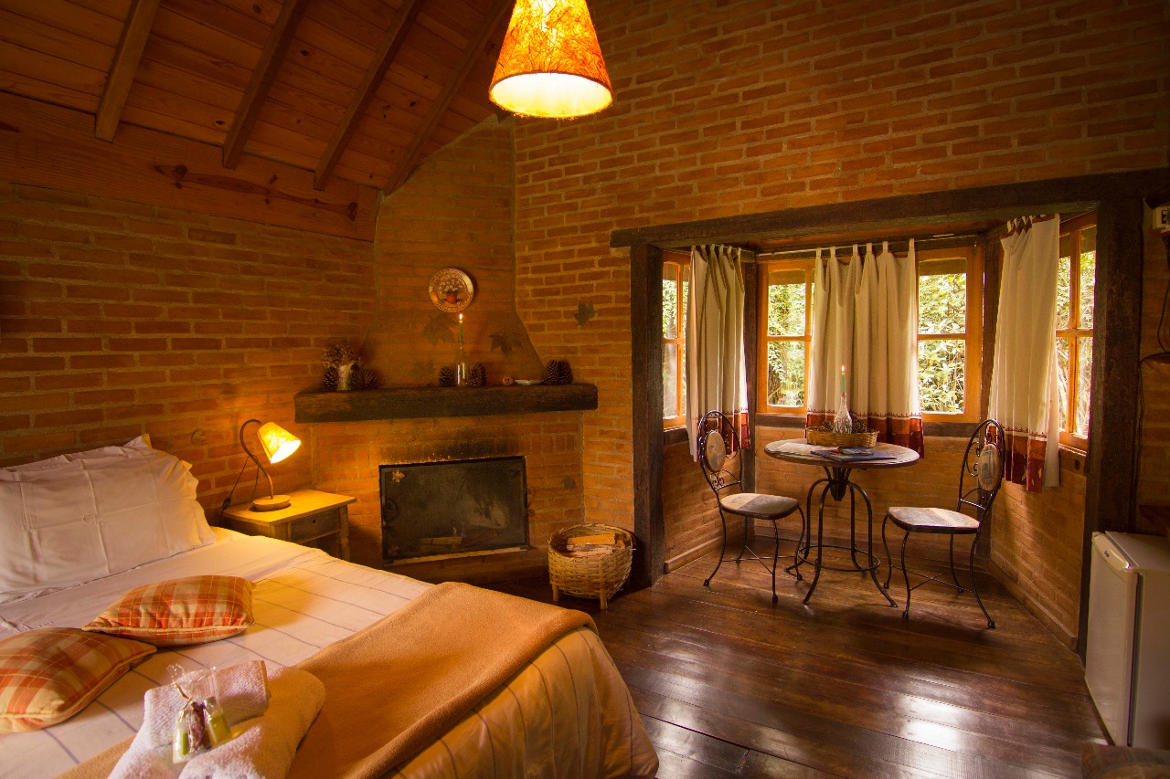 Quarto do Chalé Manacá com o abajur aceso, a lareira e a mesa junto à janela