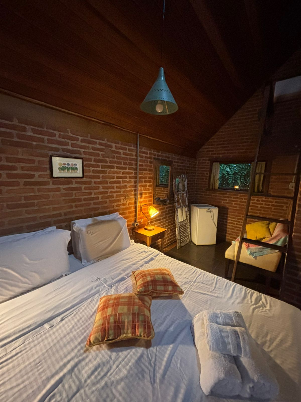 Cama do Platanus com almofadas xadrez e toalhas, sob a luminária de metal